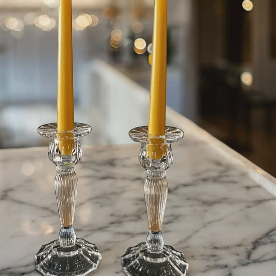 2 yellow beeswax candles in glass candleholders sat on a marble white top with the white kitchen in the background and blurred fairy lights in background. Camera moving from side to side.