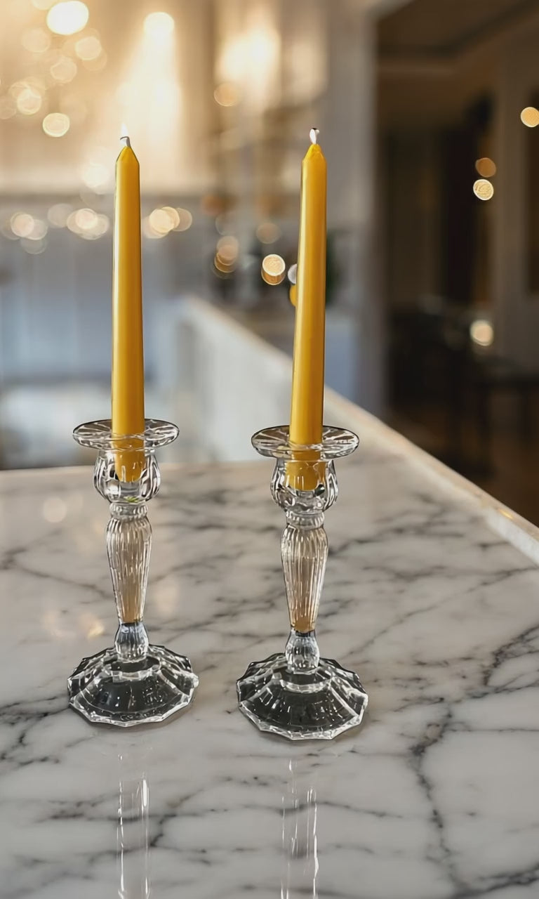 2 yellow beeswax candles in glass candleholders sat on a marble white top with the white kitchen in the background and blurred fairy lights in background. Camera moving from side to side.